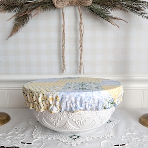 May include: A white ceramic bowl with a floral design is covered with a patchwork fabric bowl cover. The cover features yellow, blue, and white floral patterns. The bowl sits on a white lace doily, with a decorative garland in the background.