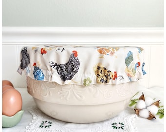 May include: A fabric bowl cover with an elasticized ruffled edge featuring a repeating pattern of colorful chickens on a cream background. The cover is placed over a large, light-colored ceramic mixing bowl with embossed floral details.