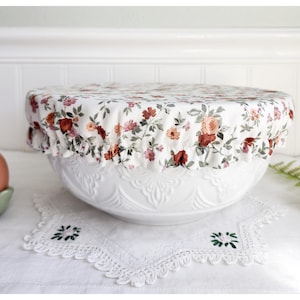 May include: A white ceramic bowl with a floral patterned fabric cover. The bowl has a decorative design and is sitting on a white surface with a lace doily.