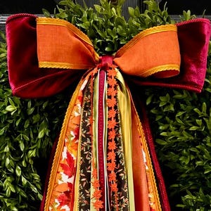 May include: A round green boxwood wreath adorned with a large multi-layered bow featuring velvet and satin ribbons in shades of burgundy, orange, and gold. Long cascading ribbon streamers hang down, displaying autumn leaf patterns and solid colors.