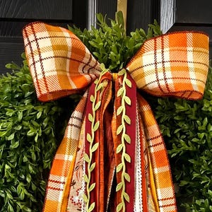 May include: A decorative wreath bow featuring a large orange and white plaid bow at the top with long, cascading ribbons in various textures and patterns, including autumn leaf prints, solid burgundy, orange, and green vine-like trim.