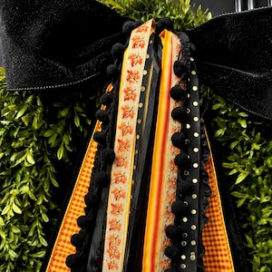 May include: A decorative bow featuring a large black velvet top, layered with multiple hanging ribbons in orange gingham, black satin, tan with orange autumn leaf prints, and black with gold polka dots, accented by black pom-pom trim.