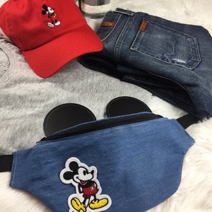 May include: A red baseball cap with a Mickey Mouse embroidered design, a denim fanny pack with Mickey Mouse patch, blue jeans, and gray athletic shoes. The items are arranged on a white fluffy surface.