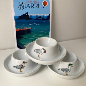 Peut inclure: Trois supports à œufs en céramique blanche avec des motifs de mouettes. Chaque support présente une mouette différente, l'une avec un objet violet dans son bec, l'autre avec un bec jaune et la dernière avec un chapeau vert.