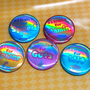 Könnte beinhalten: Fünf runde, holografische Ansteckbuttons mit dem Text I train for Speed, I train for Stamina, I train for Power, I train for Guts und I train for Wit. Jeder Button zeigt ein einzigartiges Symbol, darunter ein Bizeps, eine Flamme und ein Doktorhut.