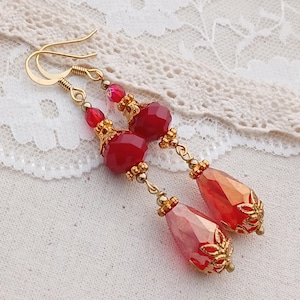 May include: A pair of long, dangling earrings with red glass beads and gold accents. The earrings have a teardrop shape and are accented with gold filigree.