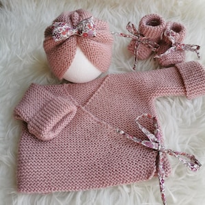 Puede incluir: Un conjunto de punto para bebé en color rosa empolvado que consta de una chaqueta cruzada de manga larga, un gorro a juego estilo turbante y un par de patucos. La chaqueta y el gorro están decorados con lazos de tela con estampado floral.
