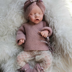 Könnte beinhalten: Eine Puppe trägt ein altrosa gestricktes Wickeloberteil mit langen Ärmeln, dazu passende gestrickte Schühchen mit Blumenband, einen passenden Strickturban mit Blumenschleife und eine cremefarbene Spitzenleggings mit zartem Lochmuster.