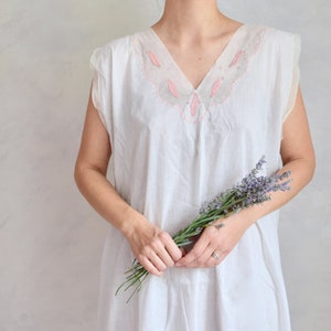 May include: A white sleeveless dress with pink embroidery around the neckline. The dress is made of a soft, lightweight fabric and has a relaxed fit. A bouquet of lavender flowers is held in the hands of the person wearing the dress.