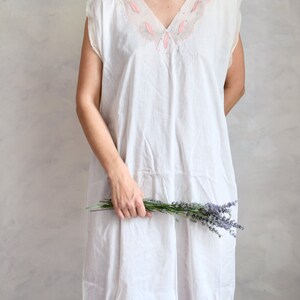 May include: A white sleeveless dress with a pink embroidered neckline. The dress is made of a lightweight fabric and has a relaxed fit. The model is holding a bouquet of lavender.