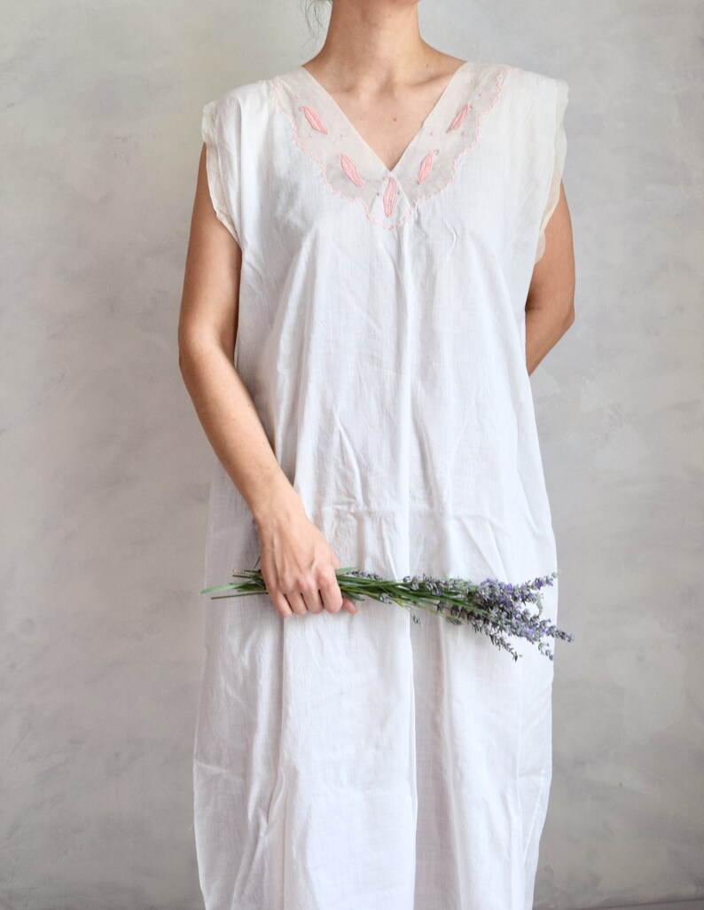 May include: A white sleeveless dress with a pink embroidered neckline. The dress is made of a lightweight fabric and has a relaxed fit. The model is holding a bouquet of lavender.