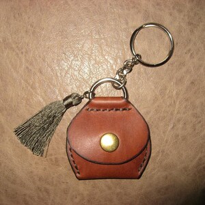 May include: A brown leather keychain with a small purse-shaped charm and a tassel. The keychain has a silver key ring and a gold button.