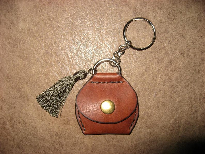 May include: A brown leather keychain with a small purse-shaped charm and a tassel. The keychain has a silver key ring and a gold button.