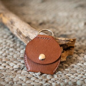 May include: A brown leather key chain with a small pocket and a silver ring. The keychain is lying on a textured surface.
