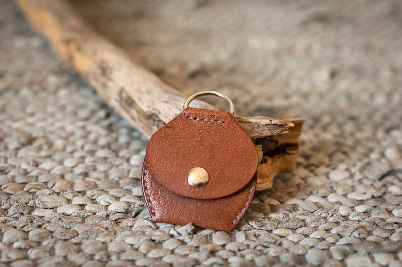 May include: A brown leather key chain with a small pocket and a silver ring. The keychain is lying on a textured surface.