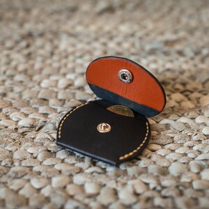 May include: A small, round, black leather coin purse with a snap closure. The purse is open, revealing a brown leather flap and a coin inside.