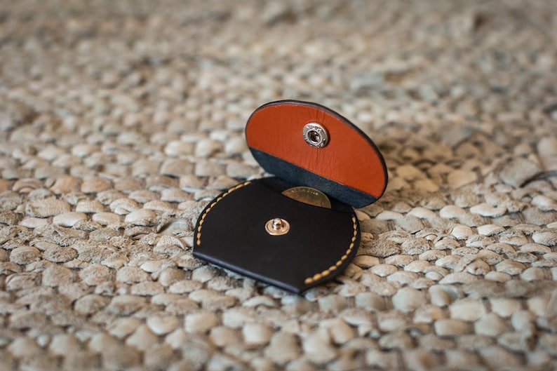 May include: A small, round, black leather coin purse with a snap closure. The purse is open, revealing a brown leather flap and a coin inside.
