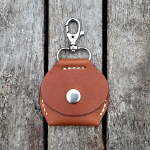 May include: A brown leather keychain case with a silver metal clasp. The case has a white stitch detail around the edges and a silver metal button on the front.