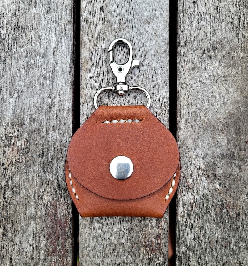 May include: A brown leather keychain case with a silver metal clasp. The case has a white stitch detail around the edges and a silver metal button on the front.