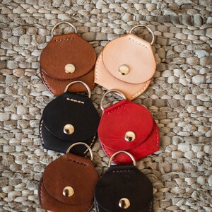 May include: Six leather keychains with a small pocket and a metal ring. The keychains are in different colors, including brown, black, red, and beige.