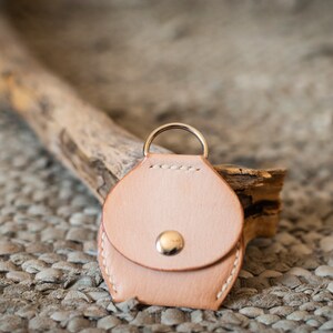 May include: A small, light brown leather keychain with a round, coin-shaped pouch and a silver ring. The keychain is sitting on a textured, brown surface.