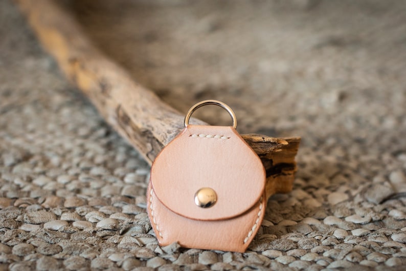 May include: A small, light brown leather keychain with a round, coin-shaped pouch and a silver ring. The keychain is sitting on a textured, brown surface.