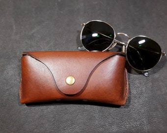 May include: A brown leather case for sunglasses. The case has a flap that closes with a button. The case is sitting on a dark gray surface.