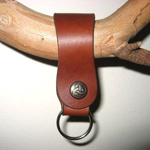 May include: A brown leather key chain with a silver metal Celtic knot design. The key chain has a ring for attaching keys.