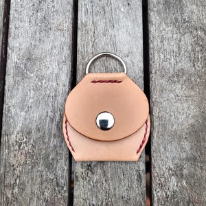 May include: A small, light brown leather coin purse with a silver ring and snap closure. The purse has red stitching around the edges.