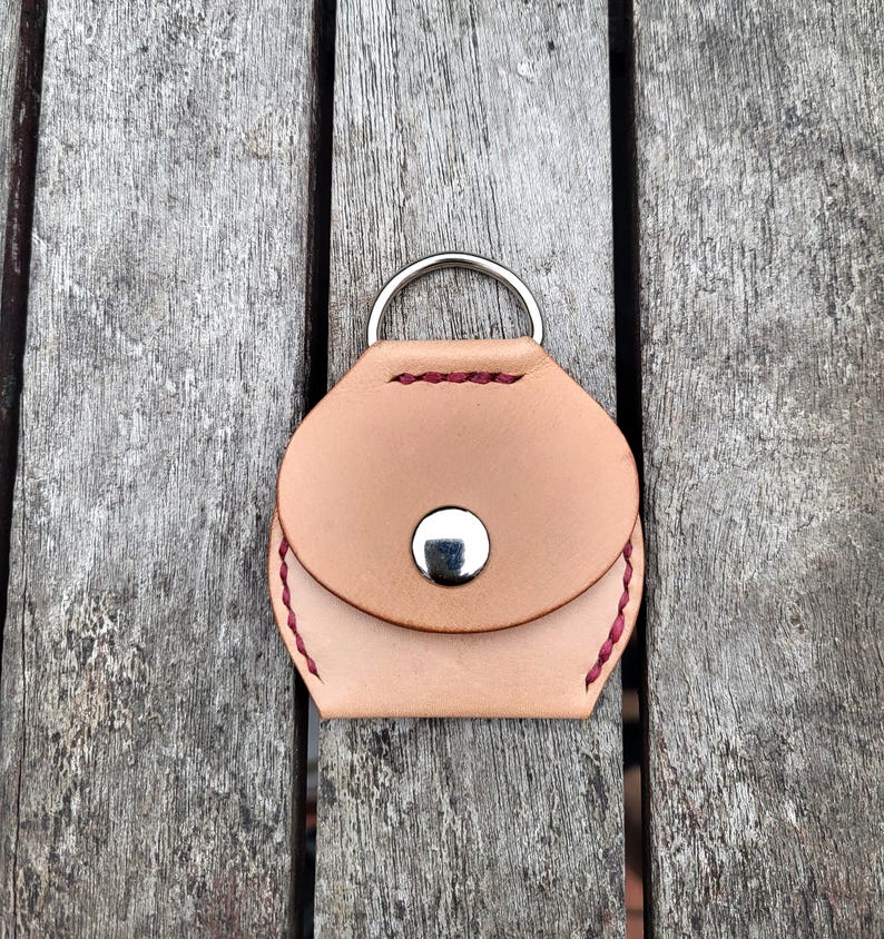 May include: A small, light brown leather coin purse with a silver ring and snap closure. The purse has red stitching around the edges.