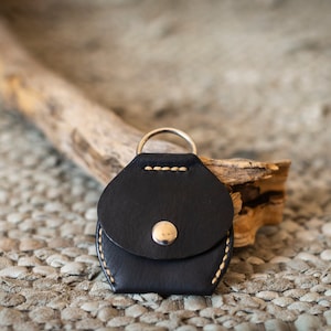 May include: A black leather key chain with a small pocket and a silver ring. The key chain is stitched with white thread and has a silver button.
