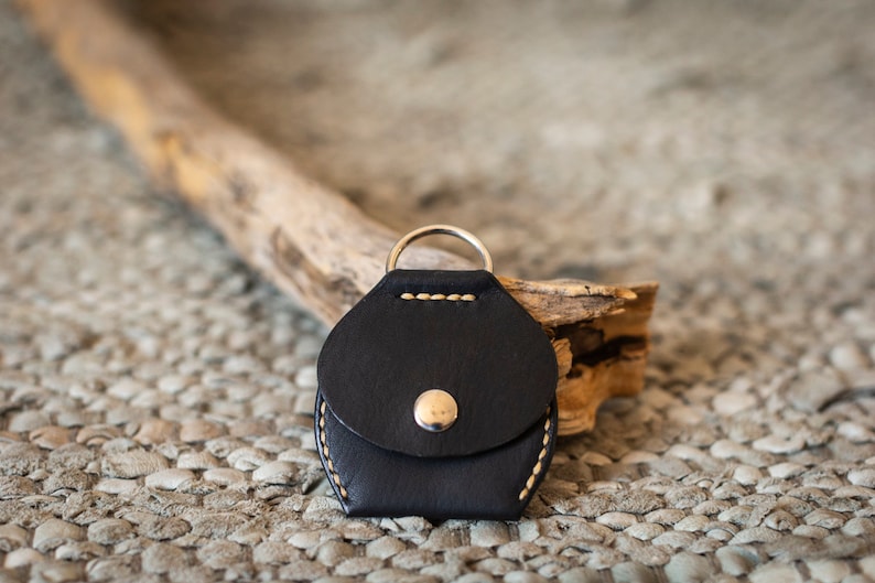 May include: A black leather key chain with a small pocket and a silver ring. The key chain is stitched with white thread and has a silver button.