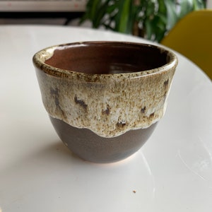 May include: A ceramic cup with a dark brown base and a speckled, light tan upper section. The interior is a rich brown. The cup has a rounded shape and a slightly flared rim. The glaze has a textured appearance.