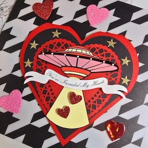 Puede incluir: Una tarjeta de San Valent&iacute;n con forma de coraz&oacute;n rojo y forma de coraz&oacute;n negro en el interior. La tarjeta presenta un OVNI rosa y rojo con el texto "You've Invaded My Heart". Corazones y estrellas brillantes est&aacute;n esparcidos alrededor de la tarjeta.