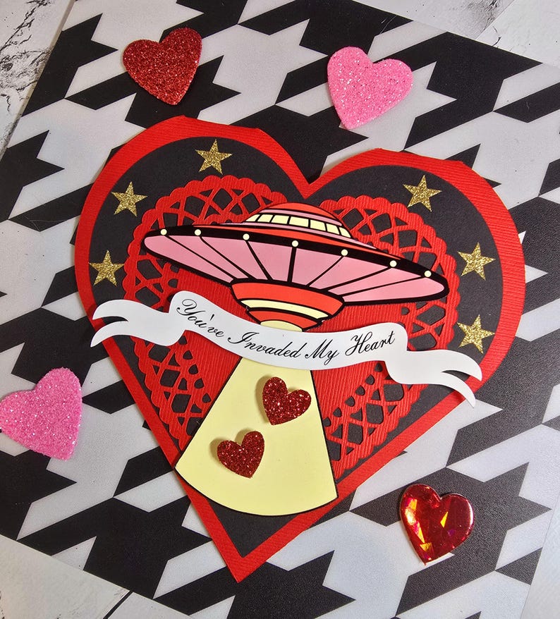 Puede incluir: Una tarjeta de San Valent&iacute;n con forma de coraz&oacute;n rojo y forma de coraz&oacute;n negro en el interior. La tarjeta presenta un OVNI rosa y rojo con el texto "You've Invaded My Heart". Corazones y estrellas brillantes est&aacute;n esparcidos alrededor de la tarjeta.