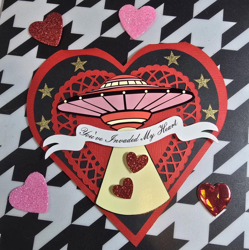 Puede incluir: Una tarjeta de San Valent&iacute;n con un coraz&oacute;n rojo, encaje negro y estrellas doradas. Un OVNI rosa y rojo est&aacute; en el centro, con el texto "You've Invaded My Heart" en una pancarta. Corazones de purpurina rosa y roja est&aacute;n dispersos.