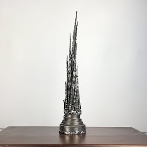 以下が含まれることがあります： 尖ったデザインの、暗いグレーの金属製の抽象彫刻。彫刻は円形、多層の台座に設置されています。彫刻は、白い背景に対して、濃い茶色の木製の表面に展示されています。