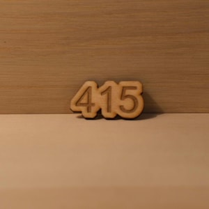 May include: A wooden cutout of the numbers 415 in a light brown color. The numbers are in a sans-serif font and are slightly raised from the surface. The background is a light brown wooden surface.