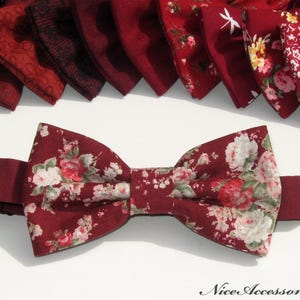 May include: A collection of bow ties in various shades of deep red, featuring patterns such as polka dots, floral prints, and solid textures. One floral bow tie is positioned in the foreground, with the text NiceAccessories4U in the corner.