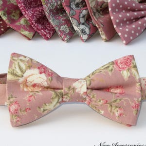 May include: A pink floral bow tie with an adjustable neckband. The bow tie displays a pattern of roses and other flowers in pink, white, and green. Several other bow ties with different patterns are visible in the background.