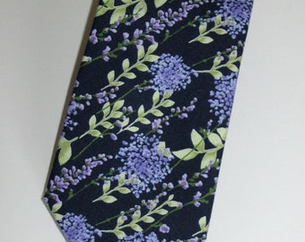 May include: A dark navy blue necktie featuring a floral pattern of lavender-colored flowers and green leaves. The tie has a classic shape and is made of a smooth fabric. The text "Nice Accessories4U" is visible.
