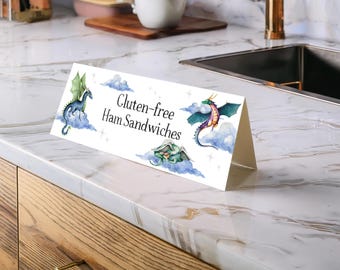 May include: A white, tent-style sign with the text "Gluten-free Ham Sandwiches" and watercolor illustrations of dragons and clouds. The sign is displayed on a marble countertop near a sink and wooden cabinets.