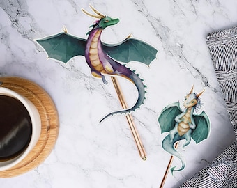 May include: Two dragon-themed pen toppers. One dragon is purple, green, and gold, the other is blue and white. The pens are gold-coloured, and the background is a marble pattern. A cup of coffee and a striped cloth are also in the image.