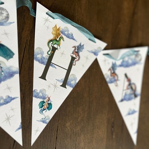 May include: Triangular banner with watercolor illustrations of dragons, clouds, and stars. The banner is strung with teal ribbon. The letter "H" is featured on one of the pennants, with a green dragon perched on top.