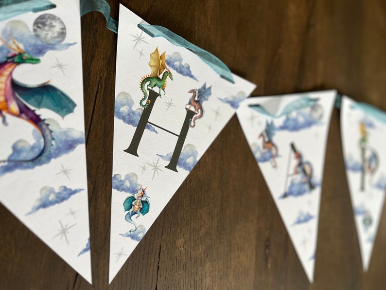 May include: Triangular banner with watercolor illustrations of dragons, clouds, and stars. The banner is strung with teal ribbon. The letter "H" is featured on one of the pennants, with a green dragon perched on top.