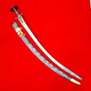 Puede incluir: Una espada de metal curvada con hoja plateada y una empuñadura ornamentada de acabado oscuro con pomo circular. Junto a ella, una vaina a juego decorada con intrincados patrones florales azules y blancos y una envoltura de tela naranja brillante.