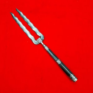 Op de afbeelding: Een tweetandige vleesvork met lange, golvende, zilverkleurige metalen tanden. Het handvat is cilindervormig, zwart van kleur en versierd met ingewikkelde zilverkleurige metalen banden en gedetailleerde gravures aan de basis en kop.