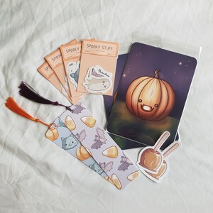 May include: A set of Halloween themed stickers featuring a ghost, a cat, a pumpkin, and a candy apple. The stickers are on a white background with a purple and orange tassel bookmark.