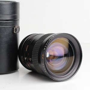 May include: A black camera lens with a silver ring and the words "MD AUTO" and "MC OKUMAR" printed on it. The lens is in a black leather case.