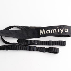 May include: A black camera neck strap made of woven fabric featuring the white text Mamiya printed on the surface. The strap includes adjustable black plastic sliders and loops at both ends for attachment to a camera body or lens equipment.